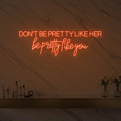 Photo of the 'DON'T BE PRETTY LIKE HER — be pretty like you' neon sign glowing in golden yellow against a styled interior wall.