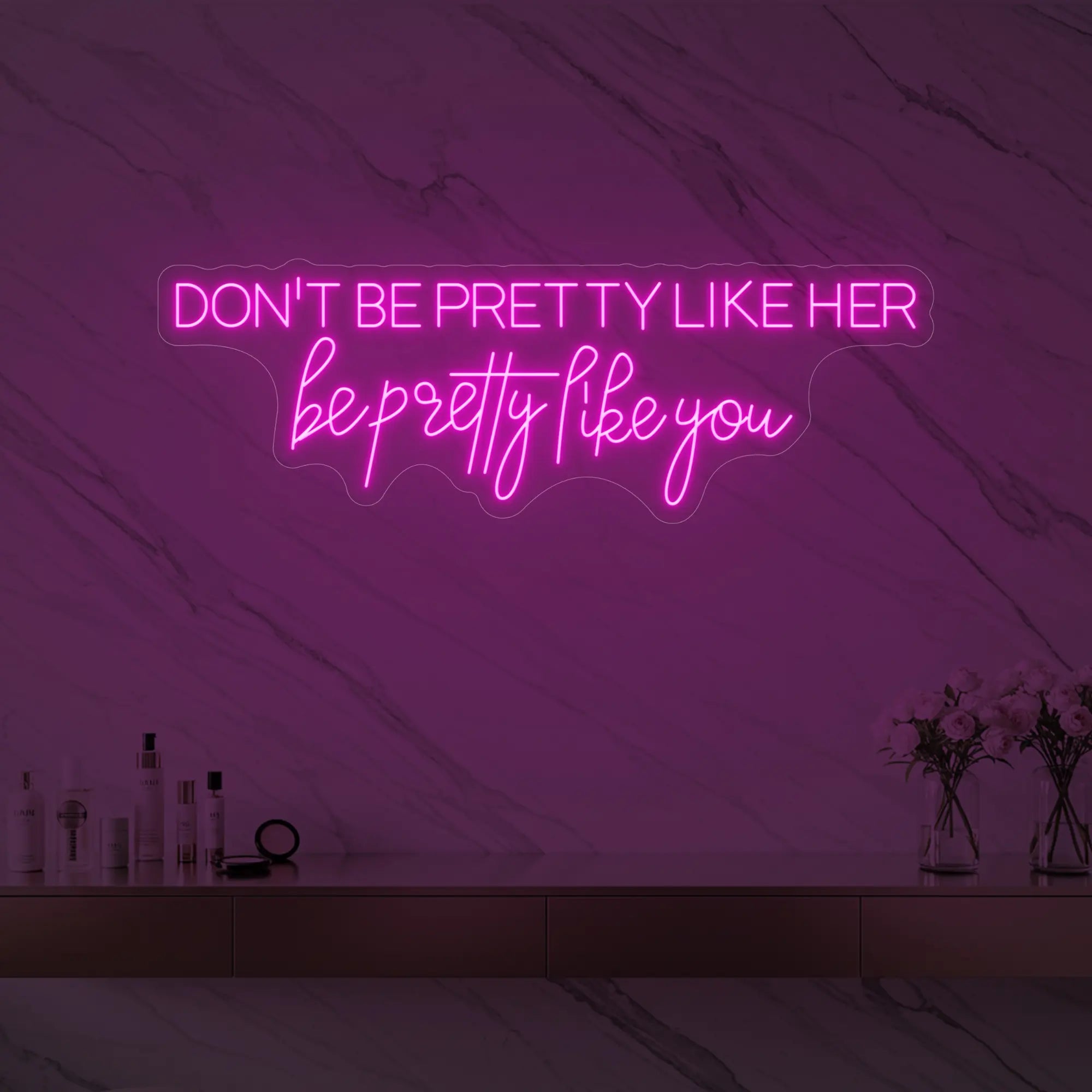 Photo of the 'DON'T BE PRETTY LIKE HER — be pretty like you' neon sign glowing in light pink against a styled interior wall.