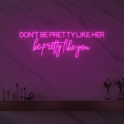 Photo of the 'DON'T BE PRETTY LIKE HER — be pretty like you' neon sign glowing in light pink against a styled interior wall.
