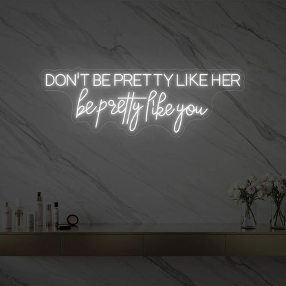 Photo of the 'DON'T BE PRETTY LIKE HER — be pretty like you' neon sign glowing in cool white against a styled interior wall.