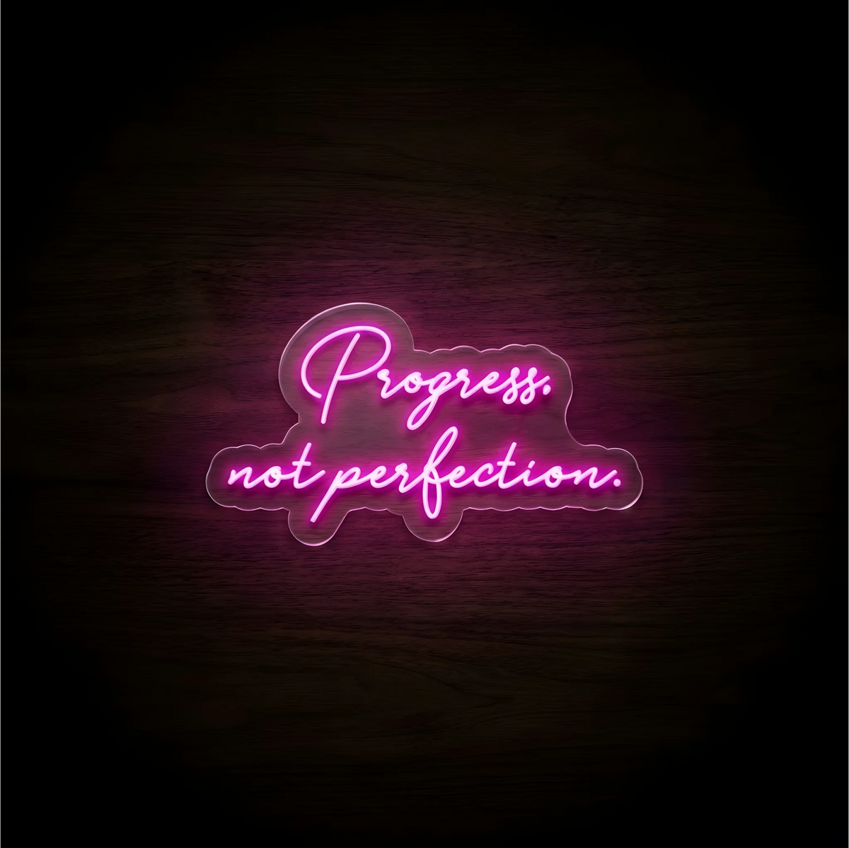 Progress. not perfection. LED Neon Sign in glowing pink script on clear acrylic against a dark wood background, ideal for home offices and creative studios.