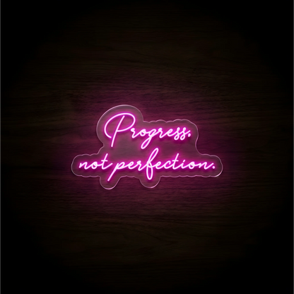 Progress. not perfection. LED Neon Sign in glowing pink script on clear acrylic against a dark wood background, ideal for home offices and creative studios.