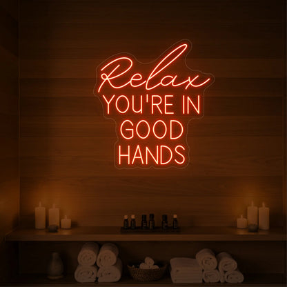 Photo of the 'Relax You're in Good Hands' neon sign glowing in golden yellow against a styled interior wall.