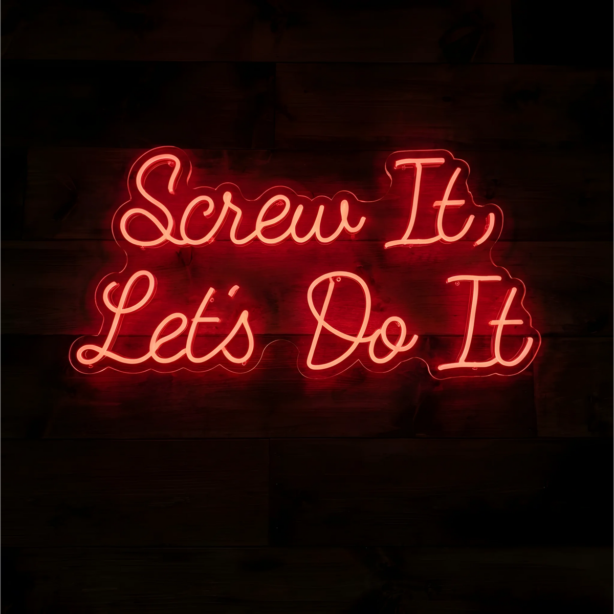 Screw It Let's Do It LED Neon Sign in glowing red-orange script lettering on clear acrylic against a dark wood background, suited to home offices and creative workspaces.