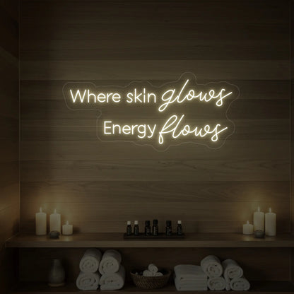 Photo of the 'Where Skin Glows, Energy Flows' neon sign glowing in warm white against a styled interior wall.