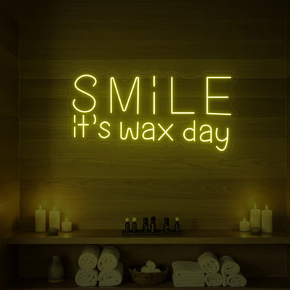 Photo of the 'Smile It’s Wax Day' neon sign glowing in lemon yellow against a styled interior wall.