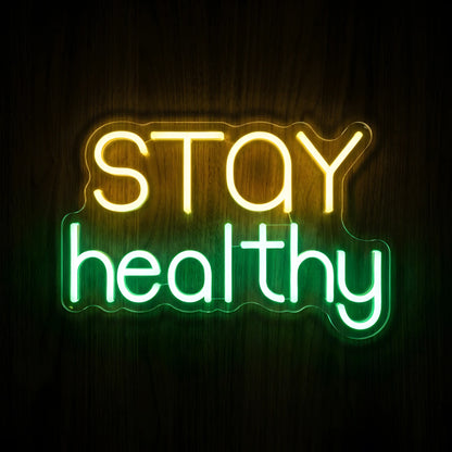 Stay Healthy LED Neon Sign featuring glowing yellow STAY lettering and bright green healthy text on clear cut-to-shape acrylic backing against a dark wooden wall.