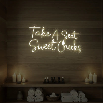 Photo of the 'Take A Seat Sweet Cheeks' neon sign glowing in cool white against a styled interior wall.