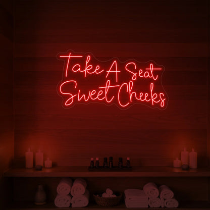 Photo of the 'Take A Seat Sweet Cheeks' neon sign glowing in red against a styled interior wall.