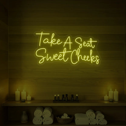 Photo of the 'Take A Seat Sweet Cheeks' neon sign glowing in lemon yellow against a styled interior wall.