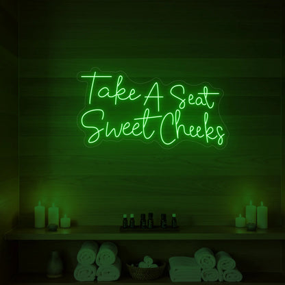 Photo of the 'Take A Seat Sweet Cheeks' neon sign glowing in green against a styled interior wall.