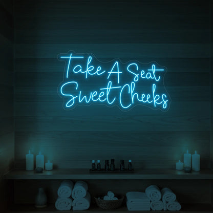 Photo of the 'Take A Seat Sweet Cheeks' neon sign glowing in ice blue against a styled interior wall.