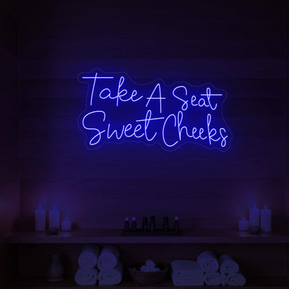 Photo of the 'Take A Seat Sweet Cheeks' neon sign glowing in deep blue against a styled interior wall.
