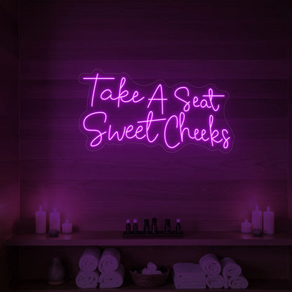 Photo of the 'Take A Seat Sweet Cheeks' neon sign glowing in deep pink against a styled interior wall.