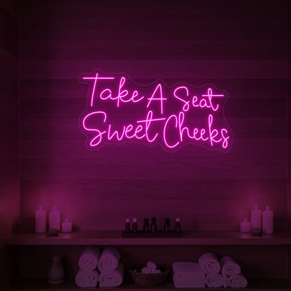 Photo of the 'Take A Seat Sweet Cheeks' neon sign glowing in light pink against a styled interior wall.