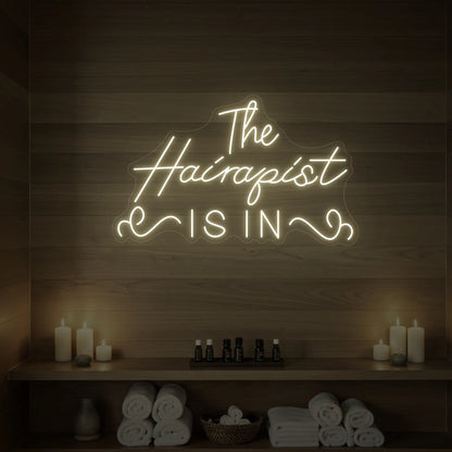 Photo of the 'The Hairapist Is In' neon sign glowing in warm white against a styled interior wall.