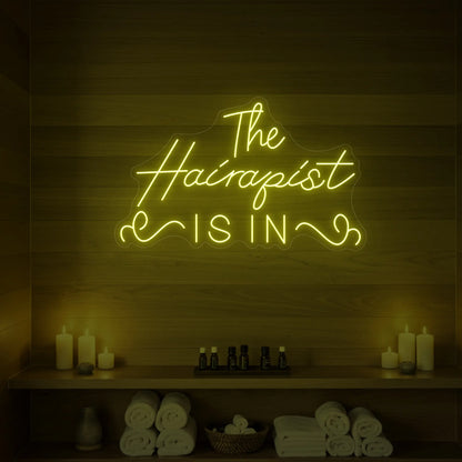 Photo of the 'The Hairapist Is In' neon sign glowing in lemon yellow against a styled interior wall.