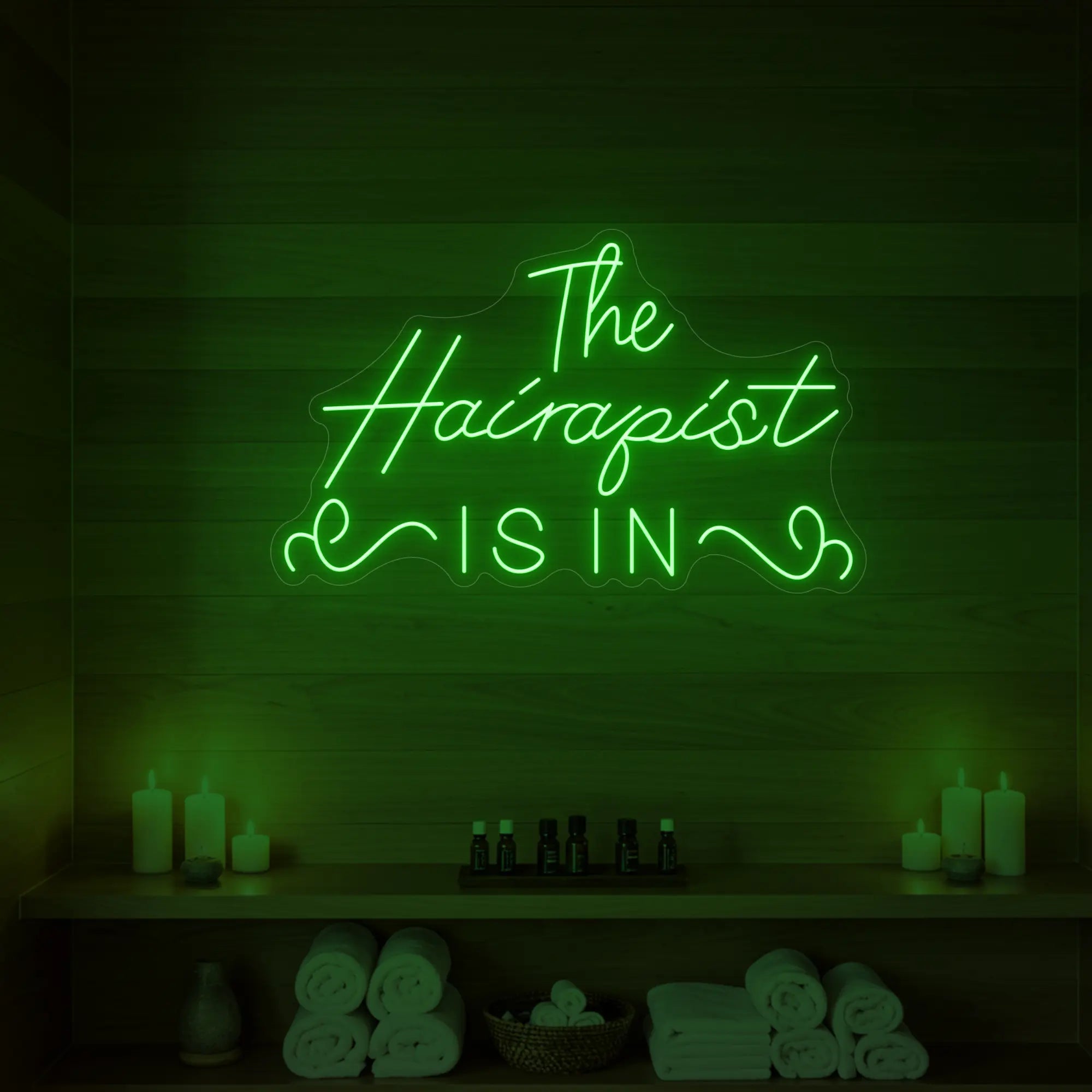 Photo of the 'The Hairapist Is In' neon sign glowing in green against a styled interior wall.