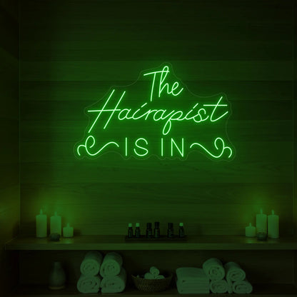 Photo of the 'The Hairapist Is In' neon sign glowing in green against a styled interior wall.