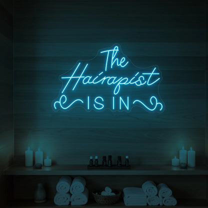 Photo of the 'The Hairapist Is In' neon sign glowing in ice blue against a styled interior wall.
