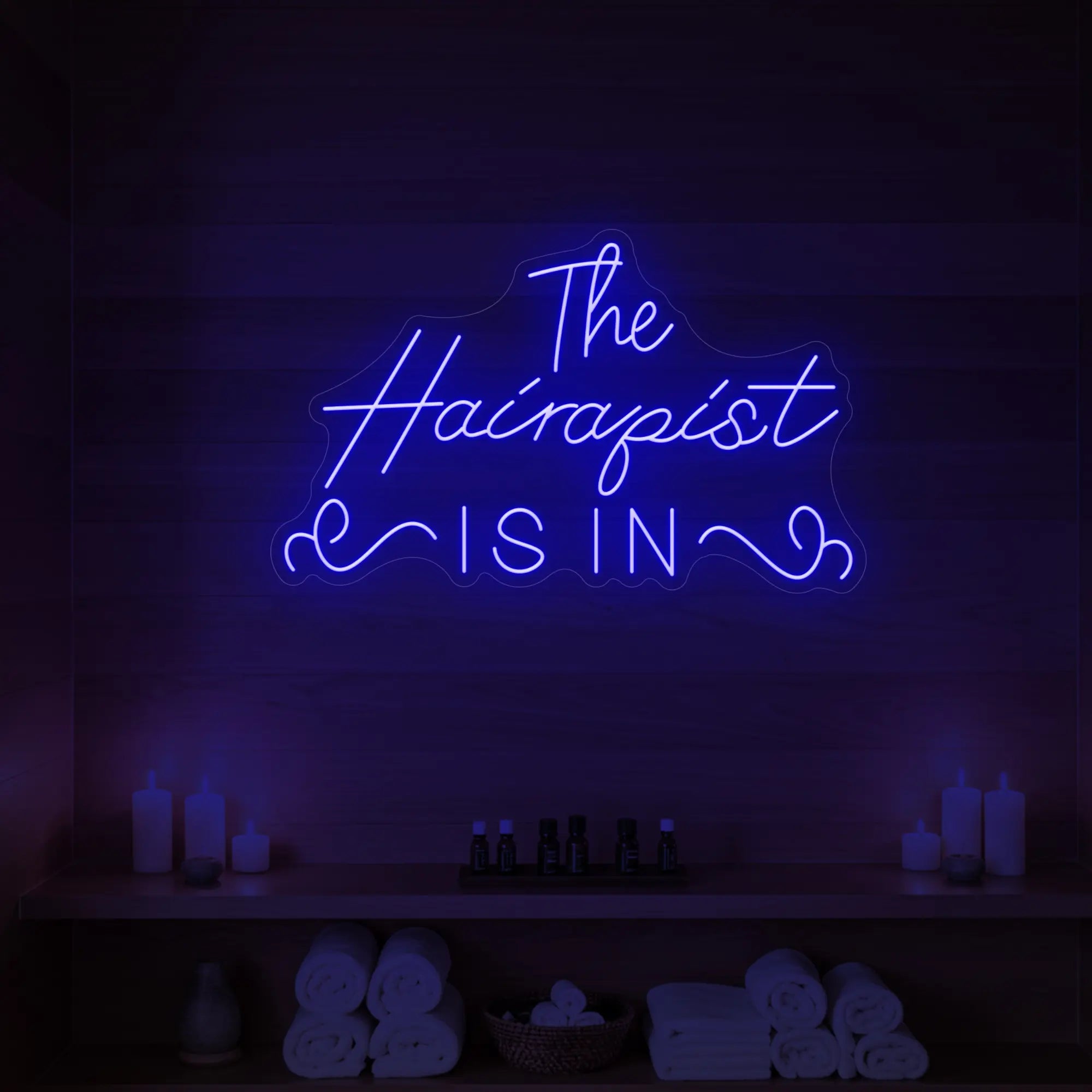 Photo of the 'The Hairapist Is In' neon sign glowing in deep blue against a styled interior wall.