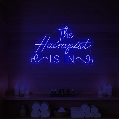 Photo of the 'The Hairapist Is In' neon sign glowing in deep blue against a styled interior wall.