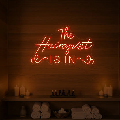 Photo of the 'The Hairapist Is In' neon sign glowing in golden yellow against a styled interior wall.