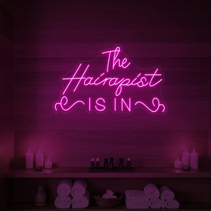 Photo of the 'The Hairapist Is In' neon sign glowing in light pink against a styled interior wall.