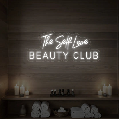 Photo of the 'The Self Love Beauty Club' neon sign glowing in cool white against a styled interior wall.