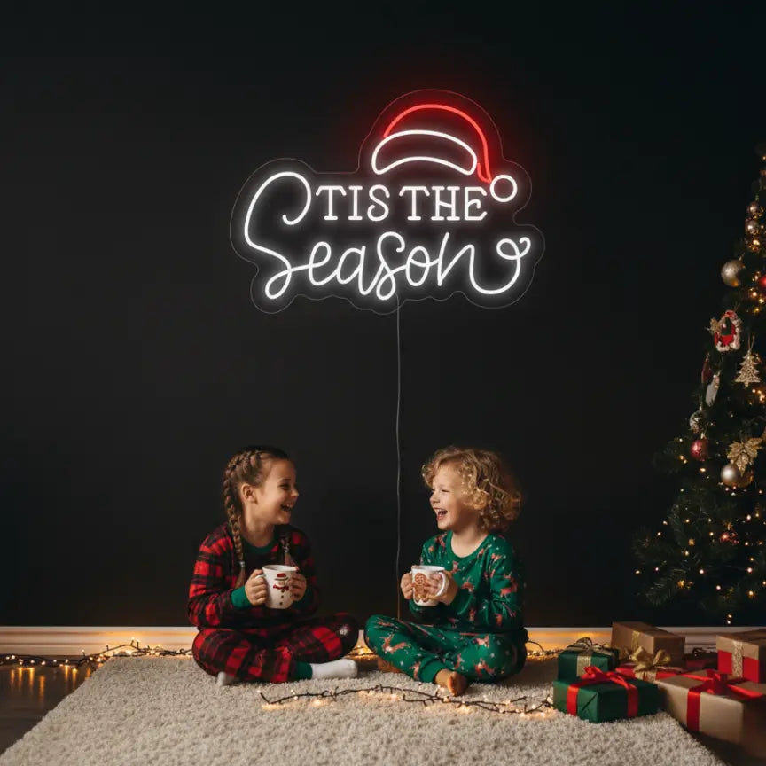 Photo of the 'Tis the Season with Santa Hat' neon sign glowing in cool white against a styled interior wall.