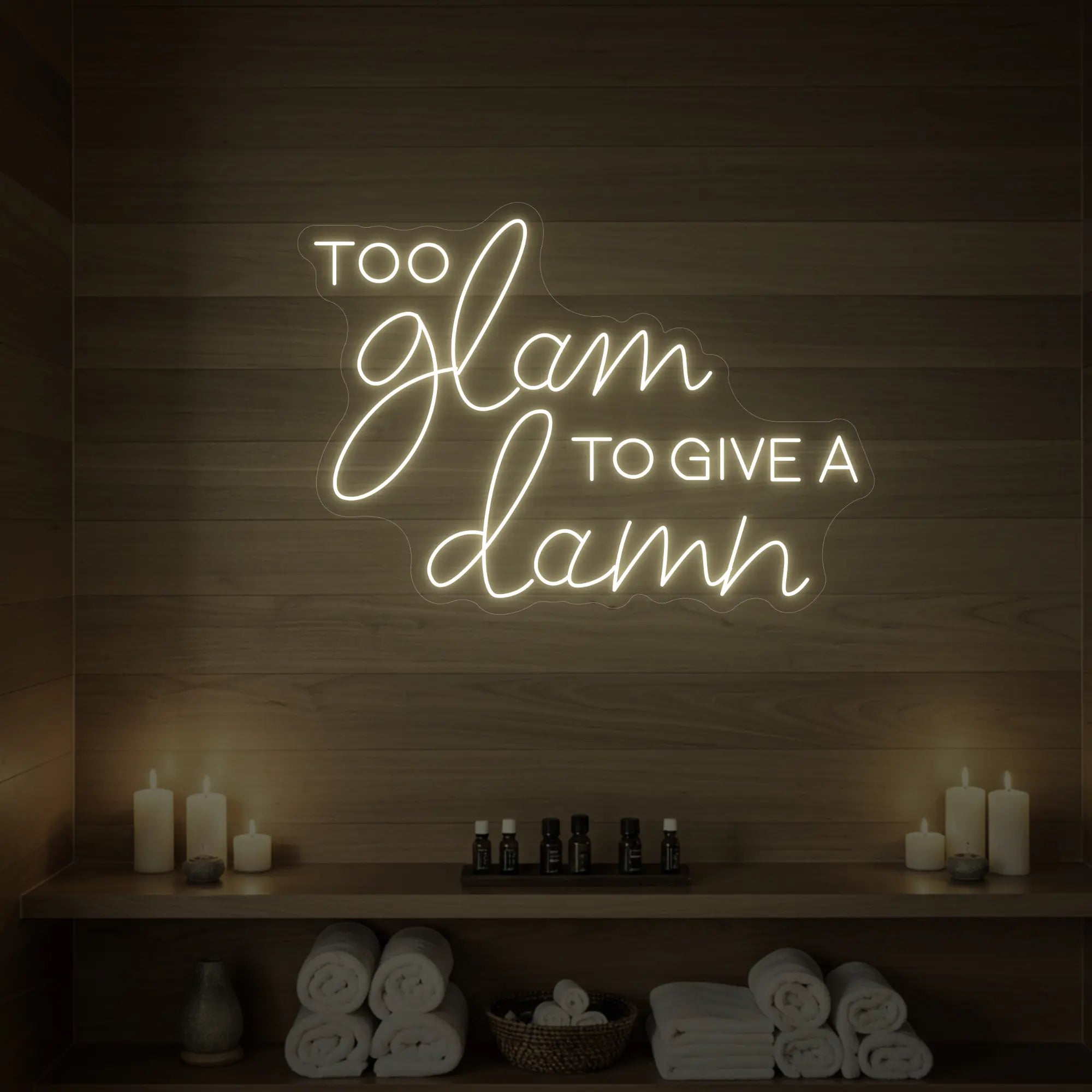 Photo of the 'TOO GLAM TO GIVE A DAMN' neon sign glowing in warm white against a styled interior wall.