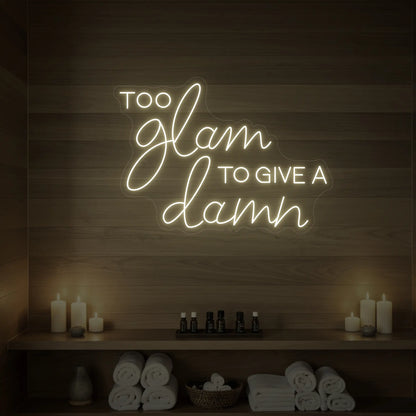 Photo of the 'TOO GLAM TO GIVE A DAMN' neon sign glowing in warm white against a styled interior wall.