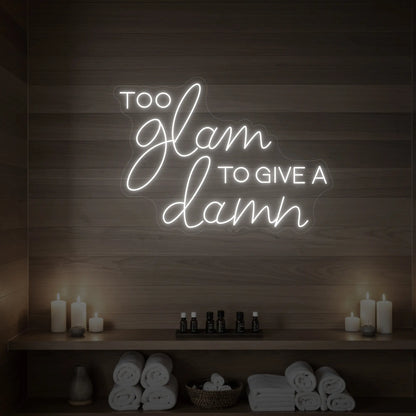 Photo of the 'TOO GLAM TO GIVE A DAMN' neon sign glowing in cool white against a styled interior wall.