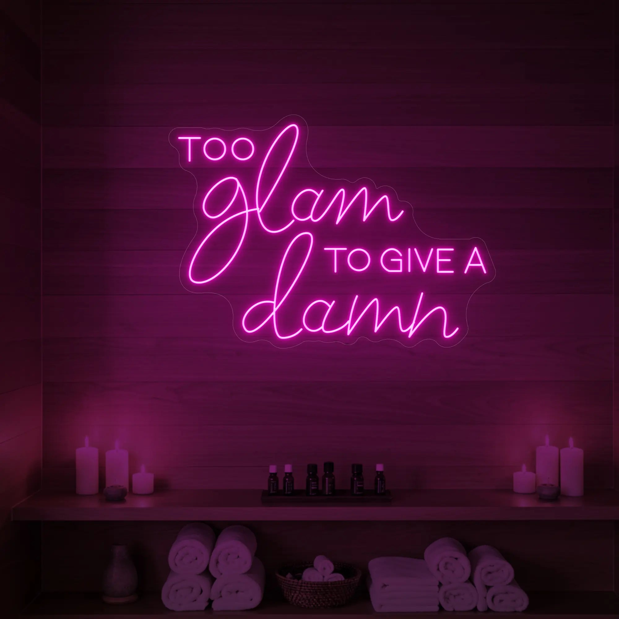 Photo of the 'TOO GLAM TO GIVE A DAMN' neon sign glowing in light pink against a styled interior wall.