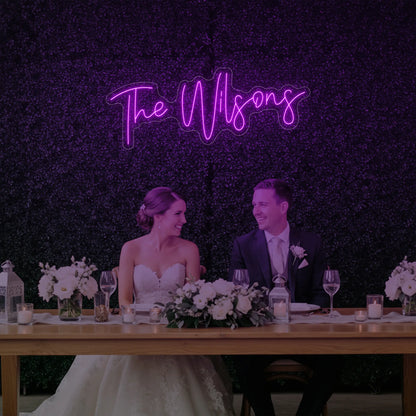 Photo of the 'Wedding Last Names Custom Neon Sign V4' neon sign glowing in deep pink against a styled interior wall.