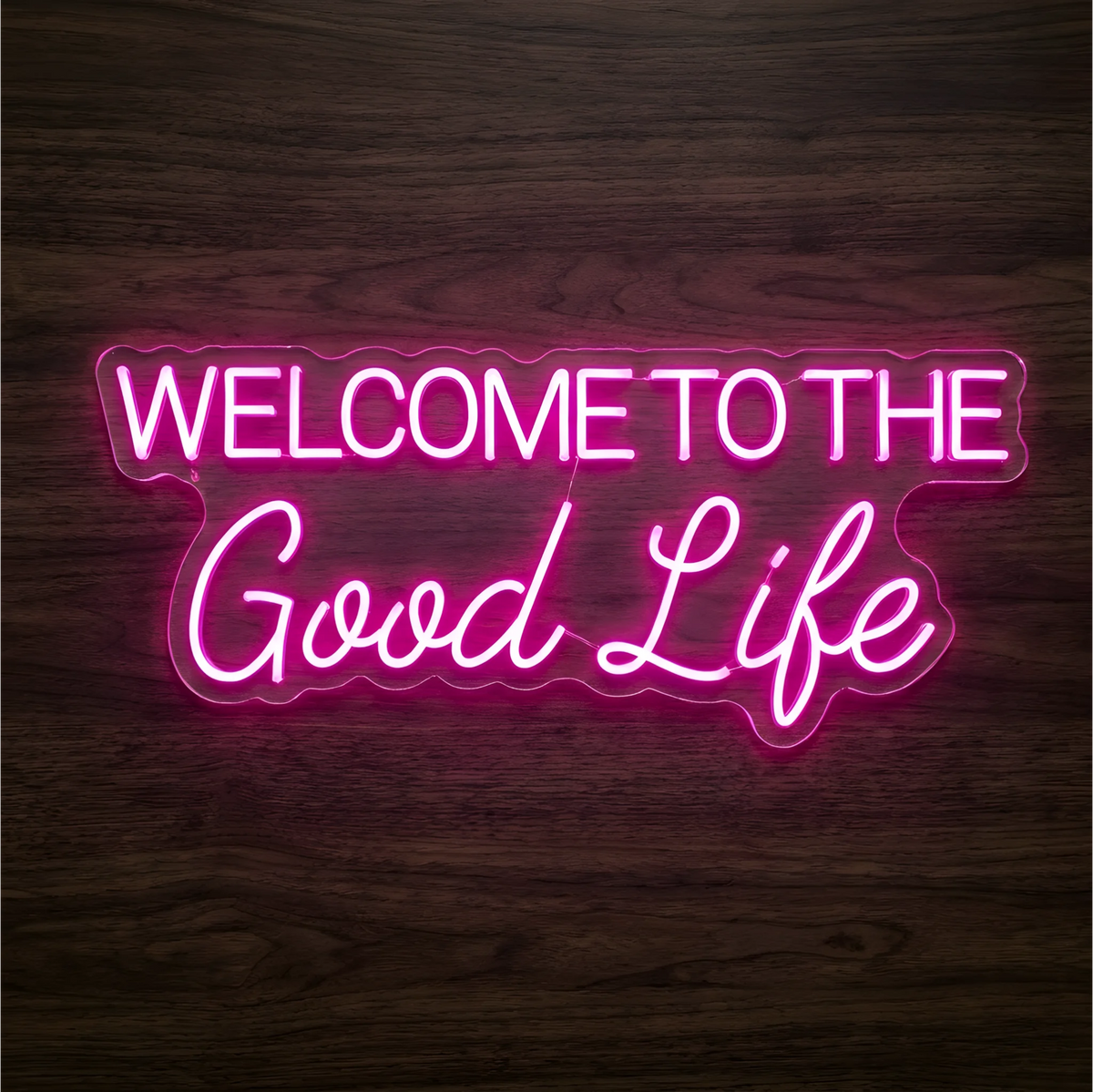 Welcome to the Good Life LED Neon Sign glowing in pink, with uppercase block lettering and flowing script on clear acrylic against a dark wood background, ideal for living rooms and boutique reception areas.