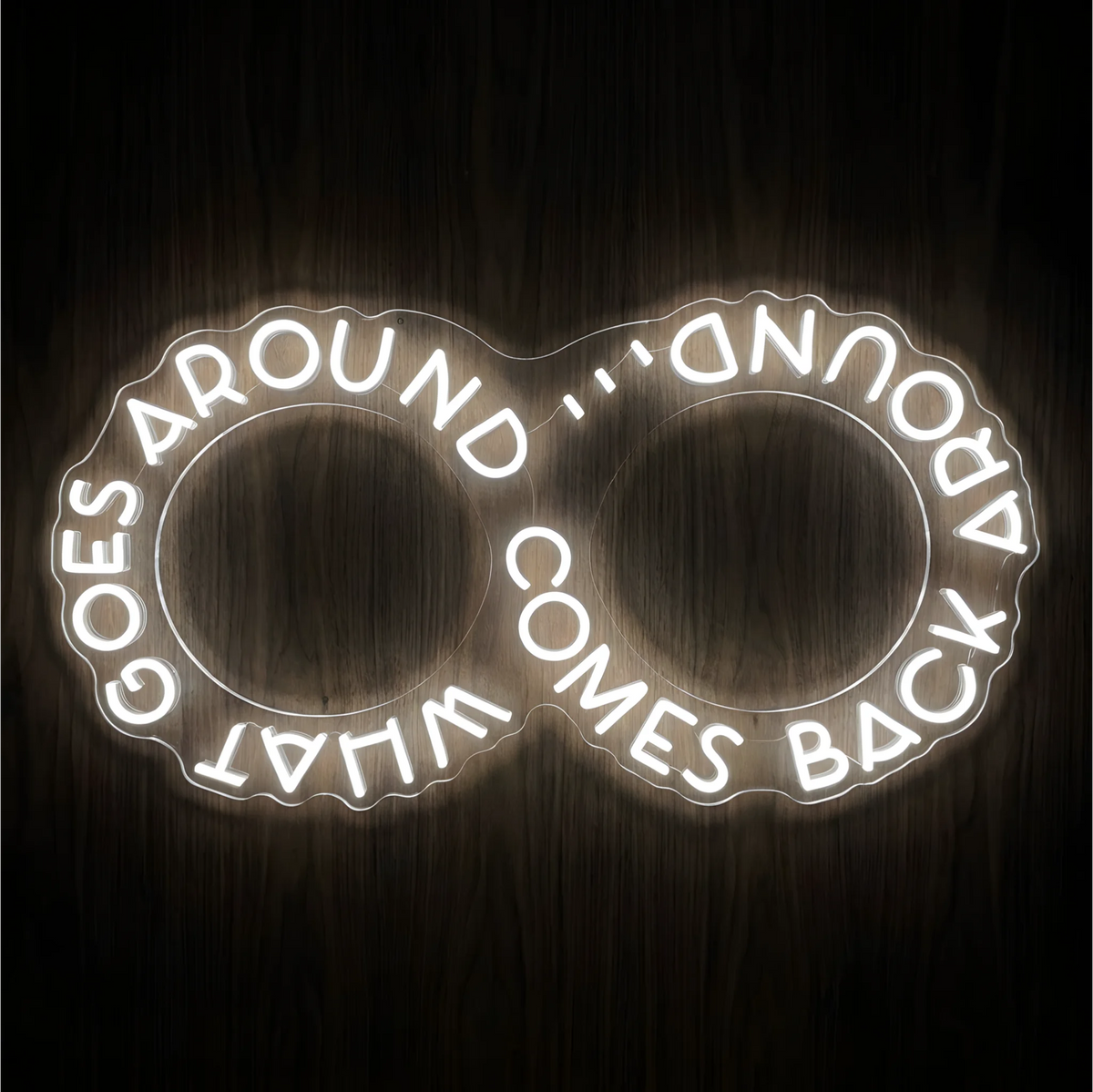 What Goes Around Comes Back Around LED Neon Sign in glowing white lettering arranged around two outlined circles on clear acrylic against a dark wood background, suited to home offices and living rooms.