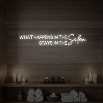 Photo of the 'WHAT HAPPENS IN THE SALON STAYS IN THE SALON' neon sign glowing in cool white against a styled interior wall.