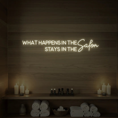 Photo of the 'WHAT HAPPENS IN THE SALON STAYS IN THE SALON' neon sign glowing in warm white against a styled interior wall.