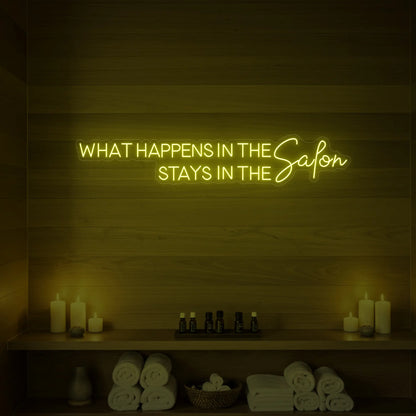 Photo of the 'WHAT HAPPENS IN THE SALON STAYS IN THE SALON' neon sign glowing in lemon yellow against a styled interior wall.