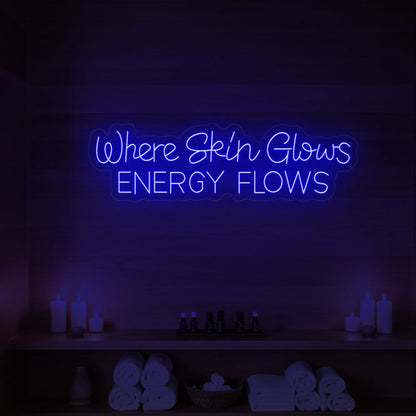 Photo of the 'Where Skin Glows, Energy Flows' neon sign glowing in deep blue against a styled interior wall.