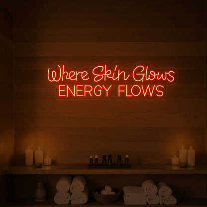 Photo of the 'Where Skin Glows, Energy Flows' neon sign glowing in golden yellow against a styled interior wall.