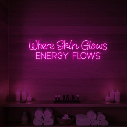 Photo of the 'Where Skin Glows, Energy Flows' neon sign glowing in light pink against a styled interior wall.