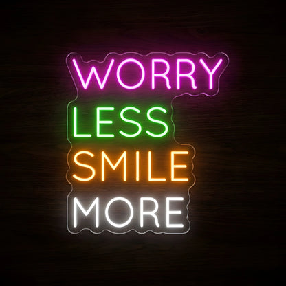 Worry Less Smile More LED neon sign featuring pink WORRY, green LESS, orange SMILE, and white MORE lettering on clear acrylic backing against a dark wooden wall.