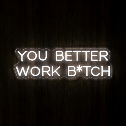“You Better Work B*TCH” LED Neon Sign glowing in bright white uppercase lettering across two lines on clear acrylic against a dark wood background, suited to home offices and creative workspaces.
