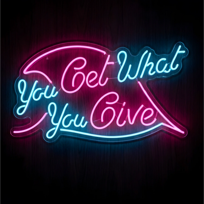 You Get What You Give LED Neon Sign in pink and cyan script with sweeping neon lines on clear acrylic against a dark wood background, suited to living rooms and creative studios.