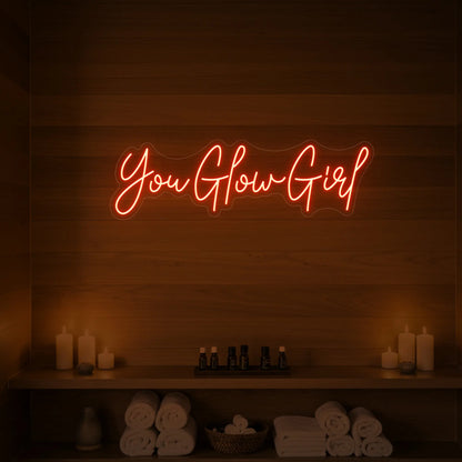 Photo of the 'You Glow Girl' neon sign glowing in golden yellow against a styled interior wall.