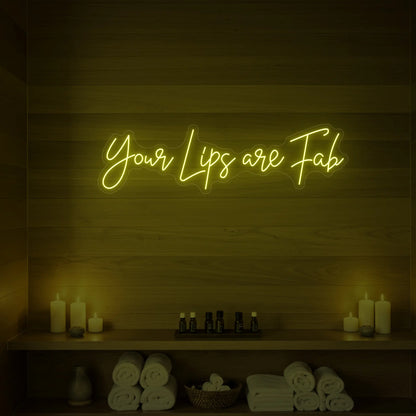 Photo of the 'Your Lips Are Fab' neon sign glowing in lemon yellow against a styled interior wall.
