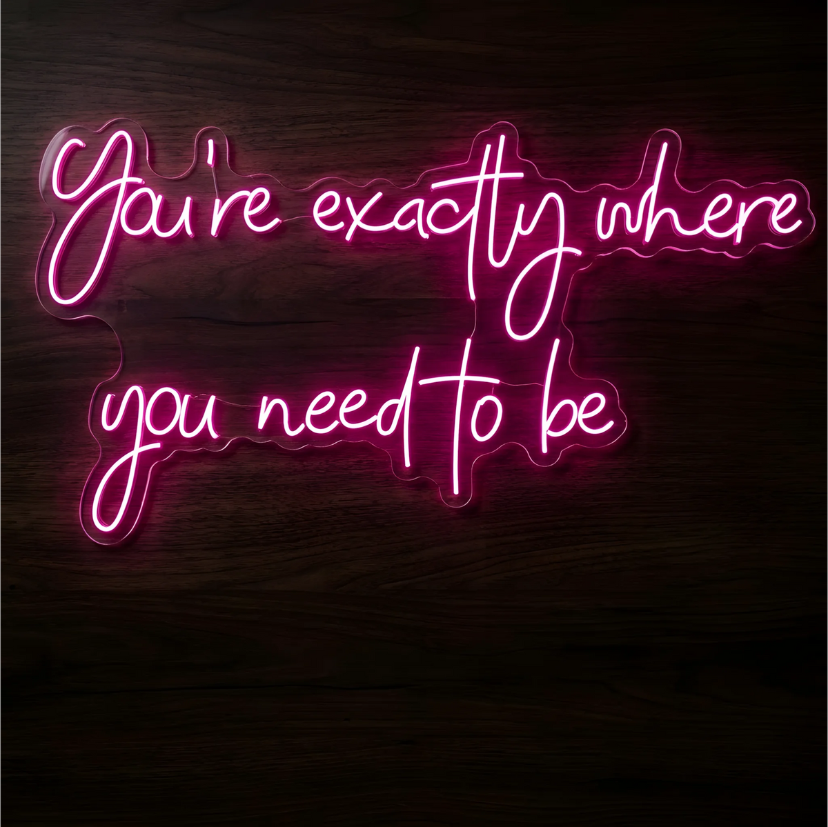 You’re Exactly Where You Need to Be LED Neon Sign in glowing pink script on clear acrylic against a dark wood background, suited to bedrooms and personal reflection spaces.